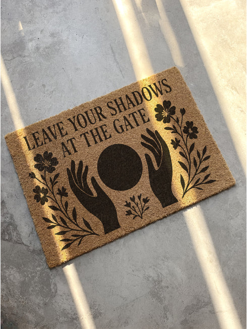 A rectangular, natural brown coir welcome mat with a textured surface, featuring an intricate, black witchy design intended for spiritual protection. The celestial and metaphysical pattern showcases a central manifestation spell circle with sacred geometry, radiating lines, and is accented with stars and crescent moons at the corners, creating a unique piece of entryway decor.